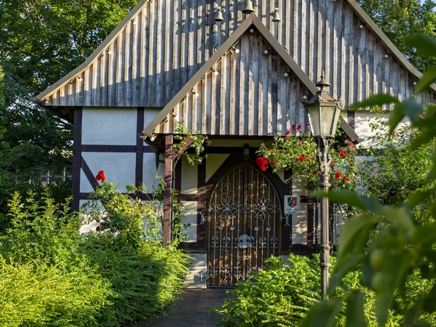 Liebharts Fachwerkdorf-Dorfkapelle Dorfkapelle mit Fachwerk und Schindeldach, umgeben von grünen Sträuchern und blühenden Rosen.