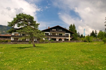 Da Nico Farchant - Außenansicht Farchanter Alm mit der Pizzeria Großes Haus mit dunklem Dach und Balkonen in weiter Entfernung, davor eine Wiese mit einem Baum, die das Haus halb verdeckt