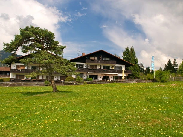 Da Nico Farchant - Außenansicht Farchanter Alm mit der Pizzeria Großes Haus mit dunklem Dach und Balkonen in weiter Entfernung, davor eine Wiese mit einem Baum, die das Haus halb verdeckt