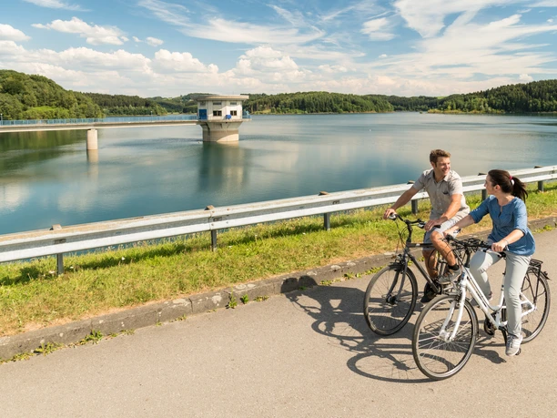 Radfahren an der Dhünntalsperre Radfahrerpaar am sonnigen Stausee, von Wäldern umgeben, unter blauem Himmel mit weißen Wolken.
