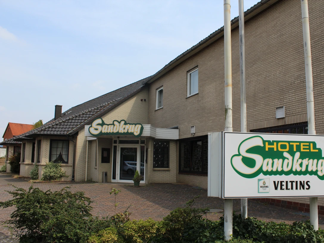 Hotel-Restaurant Sandkrug Hotel-Restaurant mit heller Backsteinfassade, grünem Logo und angrenzendem Parkplatz im Freien.Hotel-restaurant with bright brick façade, green logo and adjacent outdoor parking lot.Hotel-restaurant med lys murstensfacade, grønt logo og tilstødende udendørs parkeringsplads.Hotel-restaurant met heldere bakstenen gevel, groen logo en aangrenzende parkeerplaats.
