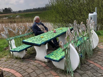 Lieblingsort Alte Staaßenbrücke-Schilfbank Eine idyllische Szene an der Alten Staaßenbrücke: Ein Mann entspannt auf einer farbenfrohen Sitzbank, umgeben von Schilfrohr-Dekorationen und frühlingshaftem Grün, während sich im Hintergrund weite Wiesen erstrecken.
