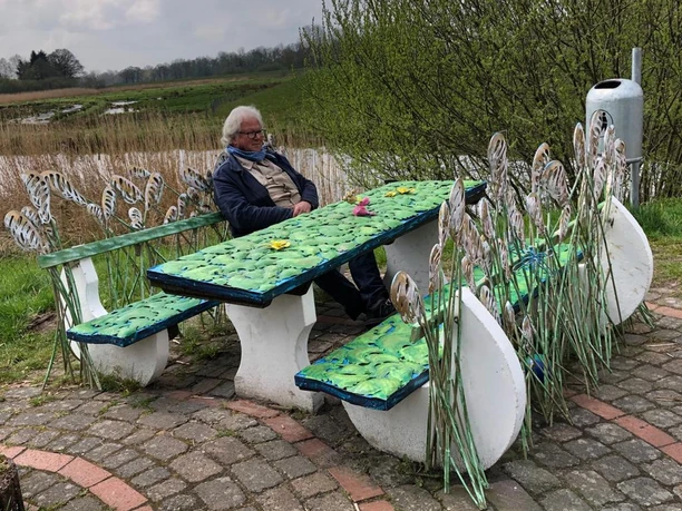 Lieblingsort Alte Staaßenbrücke-Schilfbank Eine idyllische Szene an der Alten Staaßenbrücke: Ein Mann entspannt auf einer farbenfrohen Sitzbank, umgeben von Schilfrohr-Dekorationen und frühlingshaftem Grün, während sich im Hintergrund weite Wiesen erstrecken.