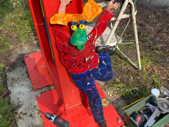 Frodewin der Frosch Ein bunter, künstlerischer Frosch mit großen, orangefarbenen Füßen und einem grünen Kopf klettert eine rote Säule am Straßenrand hoch. Werkzeug liegt um die Skulptur, was auf deren Installation hindeutet.