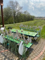 Lieblingsort am Wanderweg Das Bild zeigt einen dekorativen Rastplatz entlang eines Wanderwegs. Es umfasst kunstvoll gestaltete Tische und Bänke, verziert mit metallischen Pflanzenmotiven, die an Blätter und Gräser erinnern. Im Hintergrund erstreckt sich eine grüne Landschaft unter einem leicht bewölkten Himmel.
