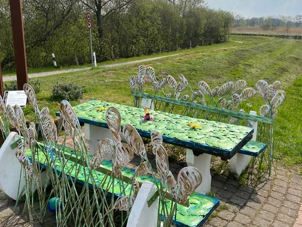 Lieblingsort am Wanderweg Das Bild zeigt einen dekorativen Rastplatz entlang eines Wanderwegs. Es umfasst kunstvoll gestaltete Tische und Bänke, verziert mit metallischen Pflanzenmotiven, die an Blätter und Gräser erinnern. Im Hintergrund erstreckt sich eine grüne Landschaft unter einem leicht bewölkten Himmel.