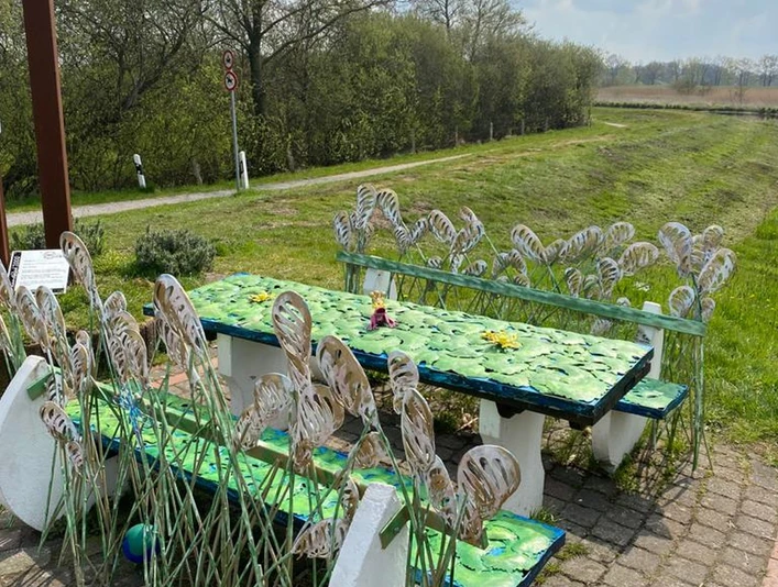 Lieblingsort am Wanderweg Das Bild zeigt einen dekorativen Rastplatz entlang eines Wanderwegs. Es umfasst kunstvoll gestaltete Tische und Bänke, verziert mit metallischen Pflanzenmotiven, die an Blätter und Gräser erinnern. Im Hintergrund erstreckt sich eine grüne Landschaft unter einem leicht bewölkten Himmel.