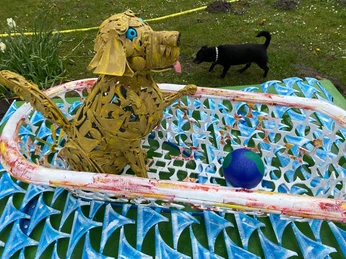 Hund und Weltkugel Ein gelber Hund aus Metall, mit herausgestreckter Zunge, sitzt in einem bunten Drahtkorb neben einem blauen Ball. Im Hintergrund ist ein schwarzer Hund auf einer Wiese sichtbar.