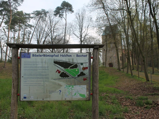 Schöpfungslehrpfad Holsten-Bexten Holztafel mit Karte des Bibelerlebnispfads Holsten-Bexten vor Kirche und Bäumen im Hintergrund