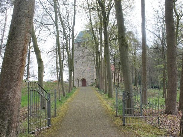 Schöpfungslehrpfad Holsten-Bexten Pflasterweg führt durch hohe Bäume zu einer alten Sandsteinkirche mit Turm und Holztor.