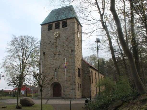 Schöpfungslehrpfad Holsten-Bexten Steinkirche mit hohem Turm und kupfernem Dach, umgeben von Bäumen am Rand eines Waldwegs