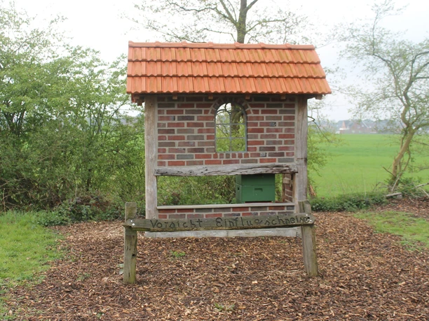Bienenlehrpfad in Salzbergen (Am Emsradweg) Backstein-Imkereihaus mit grünem Bienenkasten auf dem Naturpfad, umgeben von Bäumen und Wiese.