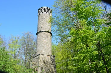 Runder steinerner Aussichtsturm im Kahlenbergwald, von üppigem Frühlingslaub umgeben, bei klarem Himmel.
