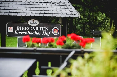 Bierenbacher Hof Biergarten Bierenbacher Hof Schild im Grünen, Blumen im Vordergrund, Sitzmöglichkeiten erkennbar.