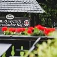 Bierenbacher Hof Biergarten Bierenbacher Hof Schild im Grünen, Blumen im Vordergrund, Sitzmöglichkeiten erkennbar.