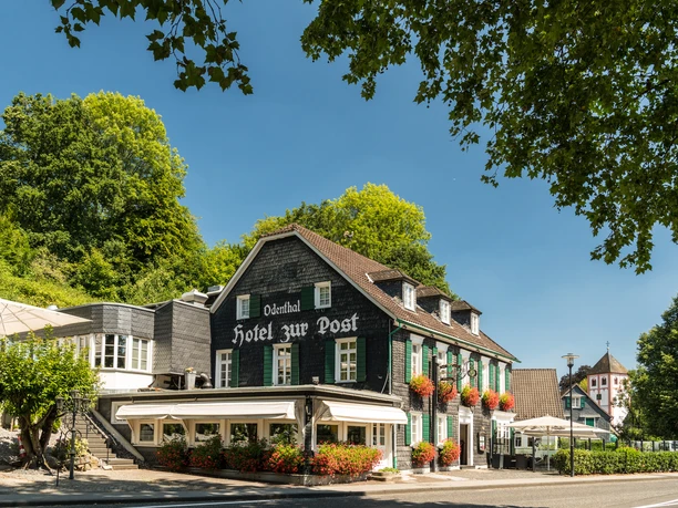 Hotel Zur Post Odenthaler Traditionshotel mit Fachwerkfassade, umgeben von grünen Bäumen, bei sonnigem Himmel.