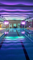 Bröltal-Bad.jpg Hallenbad mit beleuchtetem Pool und farbigen Deckenlichtern, moderne Architektur, entspannte Atmosphäre.