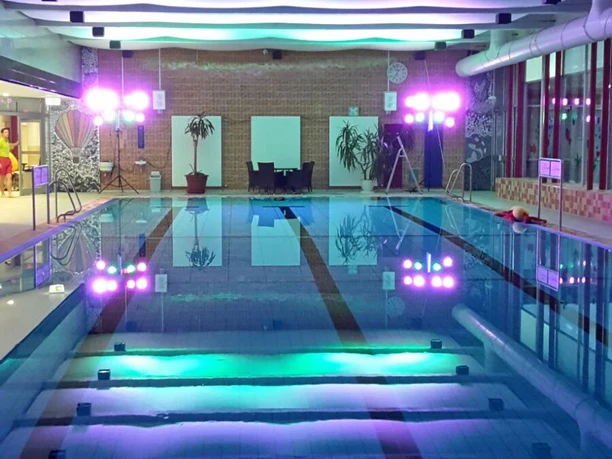 Bröltal-Bad.jpg Hallenbad mit beleuchtetem Pool und farbigen Deckenlichtern, moderne Architektur, entspannte Atmosphäre.