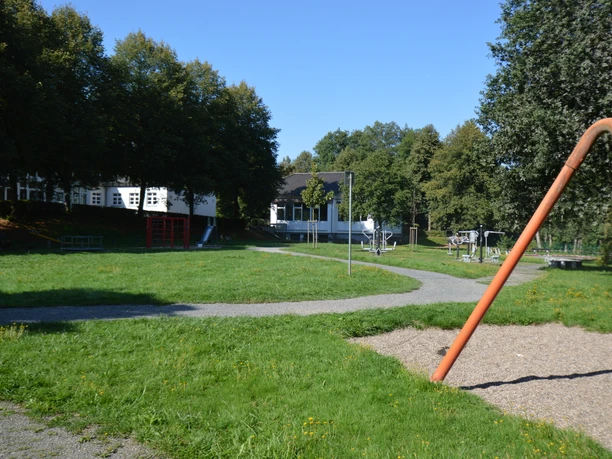 Mehrgenerationenpark Schönenberg.jpg Grüner Park mit Bäumen und Spielplatz, Schaukeln im Vordergrund, weißes Gebäude im Hintergrund.