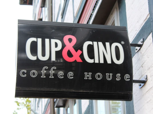 Cup & Cino Ein schwarzes Caféschild mit weißer "CUP&CINO" Aufschrift und rotem "&"-Zeichen an einer Backsteinfassade.A black café sign with white "CUP&CINO" lettering and a red "&" sign on a brick façade.Et sort caféskilt med hvide "CUP&CINO"-bogstaver og et rødt "&"-skilt på en murstensfacade.Een zwart caféteken met witte "CUP&CINO" letters en een rood "&" teken op een bakstenen gevel.