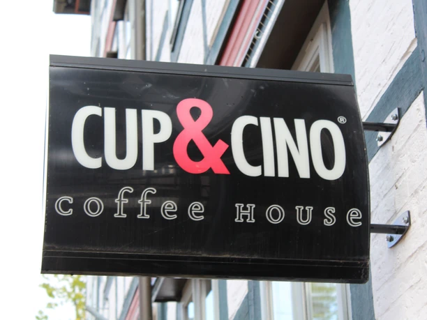 Cup & Cino Ein schwarzes Caféschild mit weißer "CUP&CINO" Aufschrift und rotem "&"-Zeichen an einer Backsteinfassade.