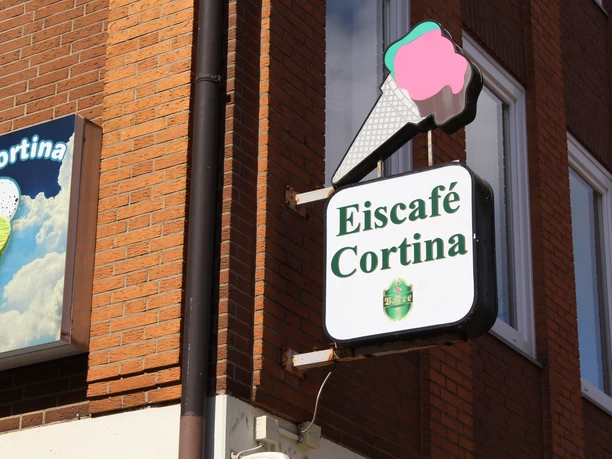 Sign for Eiscafé Cortina with ice cream waffle graphic, attached to a brick façade.