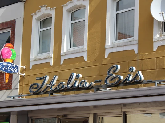 Italia Eis Original Italia Eis Café mit markantem Neon-Logo an einer gelben Fassade in urbaner Umgebung.Original Italia ice cream café with a striking neon logo on a yellow façade in an urban setting.Original Italia iscafé med et markant neonlogo på en gul facade i et urbant miljø.Origineel Italia ijscafé met een opvallend neonlogo op een gele gevel in een stedelijke omgeving.