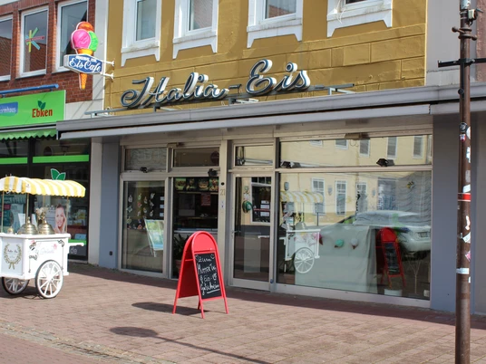 Eiscafé mit gelbem Schriftzug und Verkaufswagen vor der Glasfassade, angrenzend an ein Ladengeschäft.Ice cream café with yellow lettering and sales trolley in front of the glass façade, adjacent to a store.Iscafé med gule bogstaver og salgsvogn foran glasfacaden, ved siden af en butik.IJssalon met gele letters en verkoopwagen voor de glazen gevel, grenzend aan een winkel.