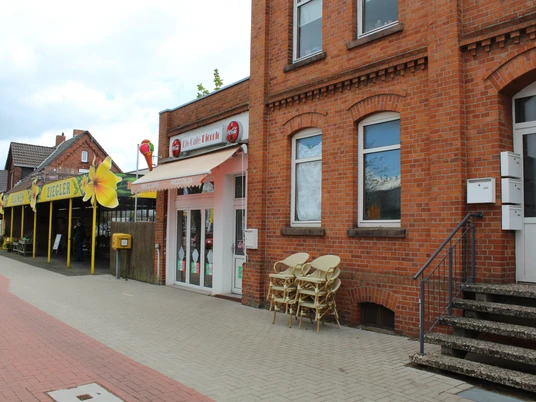 Eine rote Backsteinfassade mit Vordach, daneben Stühle; liegt an einer gepflasterten Straße.A red brick façade with a canopy, chairs next to it; located on a cobbled street.En rød murstensfacade med en baldakin, stole ved siden af; placeret på en brostensbelagt gade.Een rode bakstenen gevel met een luifel, stoelen ernaast; gelegen aan een geplaveide straat.