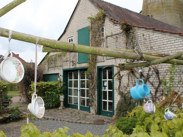 Enking´s Mühle in Emsbüren - Eingang Mühlencafé ©Emsland Tourismus GmbH, Martina Alfers.JPG Backsteinhaus mit grünen Fenstern und Türen, davor hängen bunte Tassen an Bambusstäben.