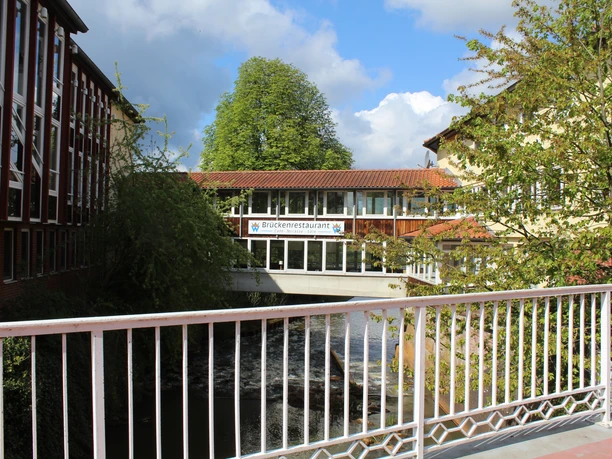 A restaurant on a pedestrian bridge connects two buildings over a river in lush nature.