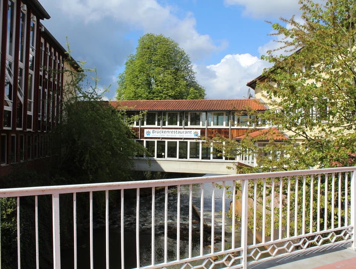 Ein Restaurant auf einer Fußgängerbrücke verbindet zwei Gebäude über einem Fluss in üppiger Natur.A restaurant on a pedestrian bridge connects two buildings over a river in lush nature.En restaurant på en fodgængerbro forbinder to bygninger over en flod i et frodigt naturområde.Een restaurant op een voetgangersbrug verbindt twee gebouwen over een rivier in een weelderige natuurlijke omgeving.