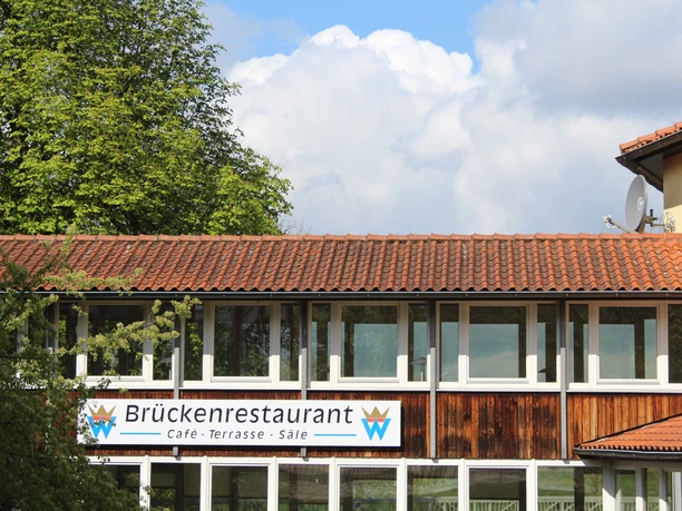 Brückenrestaurant Ein zweistöckiges Restaurant mit roten Ziegeldächern, großen Fenstern und einem blauen Himmel im Hintergrund.