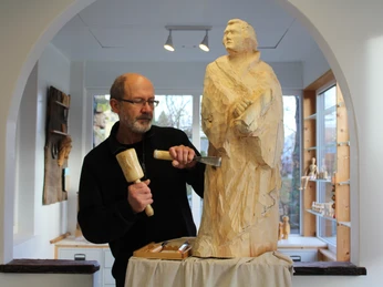 Deutsches Kreativzentrum Holz Ein Holzbildhauer arbeitet konzentriert an einer detailreichen Holzskulptur in einem hellen Raum.