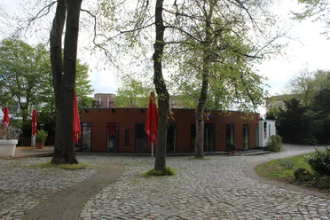 Ein kleines Backsteingebäude mit großen Fenstern, umgeben von Laubbäumen und gepflasterten Wegen.