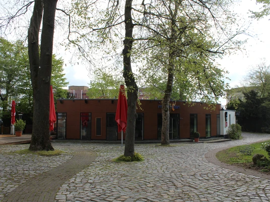 Maximillian Ein kleines Backsteingebäude mit großen Fenstern, umgeben von Laubbäumen und gepflasterten Wegen.A small brick building with large windows, surrounded by deciduous trees and paved paths.En lille murstensbygning med store vinduer, omgivet af løvtræer og asfalterede stier.Een klein bakstenen gebouw met grote ramen, omringd door loofbomen en geplaveide paden.