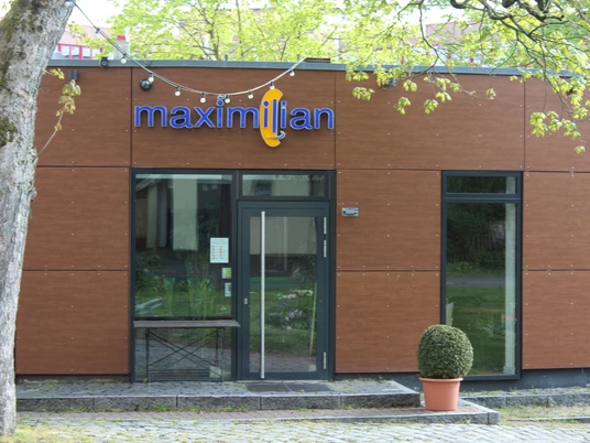 Maximillian Ein moderner Gebäudeeingang aus braunem Holz mit großen Fenstern, neben einem Baum und einem Strauch.A modern building entrance made of brown wood with large windows, next to a tree and a shrub.Et moderne indgangsparti af brunt træ med store vinduer ved siden af et træ og en busk.Een moderne ingang van bruin hout met grote ramen, naast een boom en een struik.