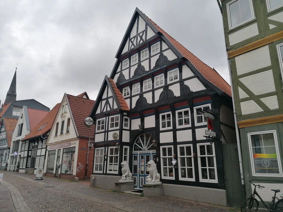 Fachwerkhaus mit roten Dachziegeln und zwei steinernen Löwen am Eingangstor, flankiert von Nachbarhäusern.