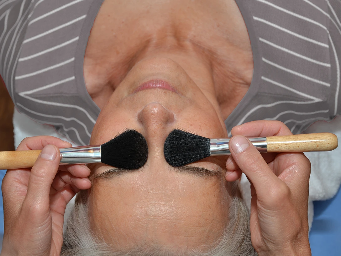 Eine Frau mit geschlossenen Augen genießt eine entspannende Behandlung mit zwei weichen Bürsten.