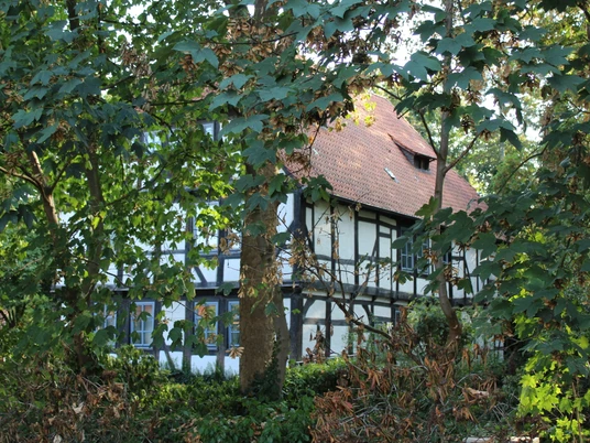 Ein historisches Fachwerkhaus mit rotem Ziegeldach, umgeben von üppigem Grün und dichten Bäumen.