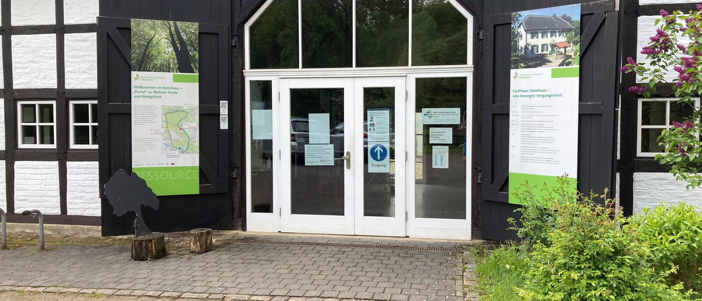 Forsthaus Steinhaus Eingang eines Fachwerkgebäudes mit weißen Wänden, großen Glastüren und informativen Schautafeln.