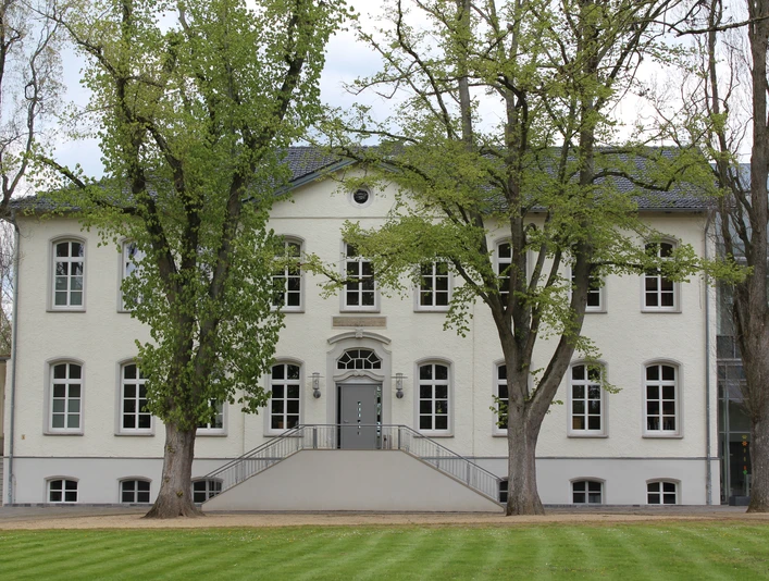 Besselsche Hof Ein weißes, zweistöckiges Gebäude mit symmetrischen Fenstern, umgeben von grünen Bäumen und Rasenflächen.