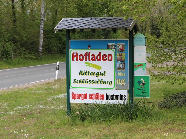 Rittergut Schlüsselburg Hinweisschild auf den Hofladen des Ritterguts Schlüsselburg, umgeben von grüner Landschaft.