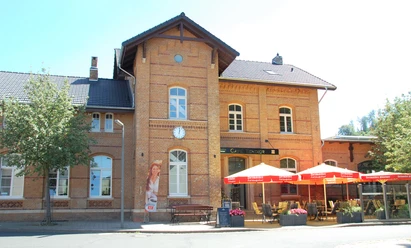 Backsteingebäude mit großen Fenstern; roter Sonnenschirm markiert das Café mit Außensitzplätzen.