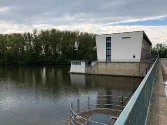 Wehr in Landesbergen mit modernem Gebäude und Metallgeländer am Fluss unter bewölktem Himmel.