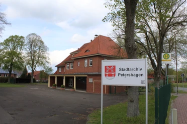 Stadtarchiv Petershagen Das Stadtarchiv Petershagen mit rotem Backsteingebäude und Bushaltestelle, umgeben von grünen Bäumen.