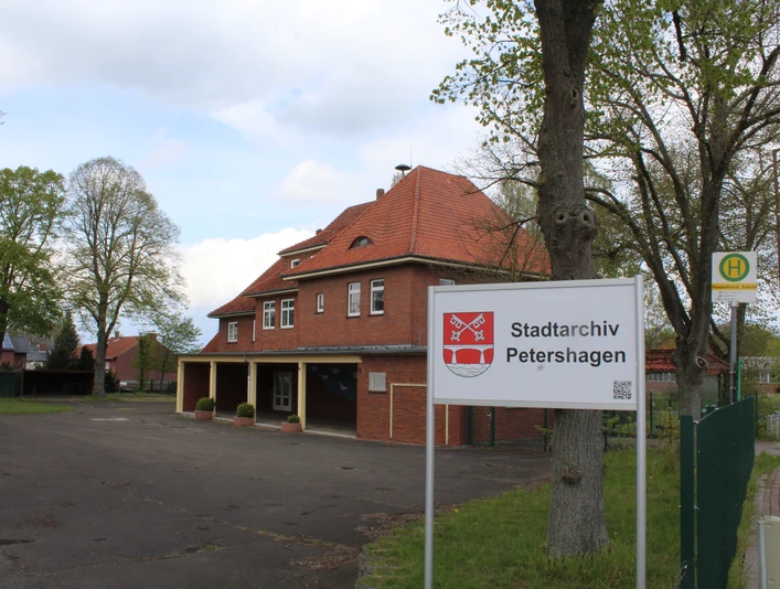 Stadtarchiv Petershagen Das Stadtarchiv Petershagen mit rotem Backsteingebäude und Bushaltestelle, umgeben von grünen Bäumen.