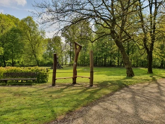 20210519_094832.jpg Holzskulpturen und eine Sitzbank am Rande einer grünen Wiese, umgeben von Bäumen im Frühling.