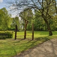 20210519_094832.jpg Holzskulpturen und eine Sitzbank am Rande einer grünen Wiese, umgeben von Bäumen im Frühling.
