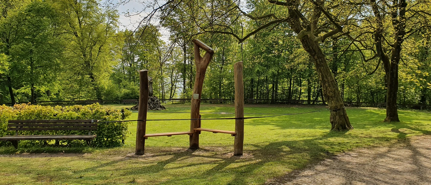 20210519_094832.jpg Holzskulpturen und eine Sitzbank am Rande einer grünen Wiese, umgeben von Bäumen im Frühling.